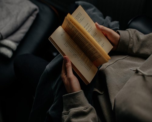 A person reading a printed book under a warm reading lamp in the evening